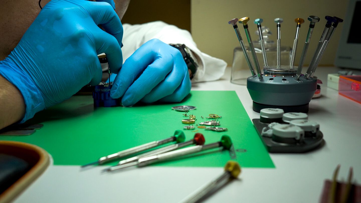 A watchmaker at his desk with movement parts and tools laid out on a clean green work surface.