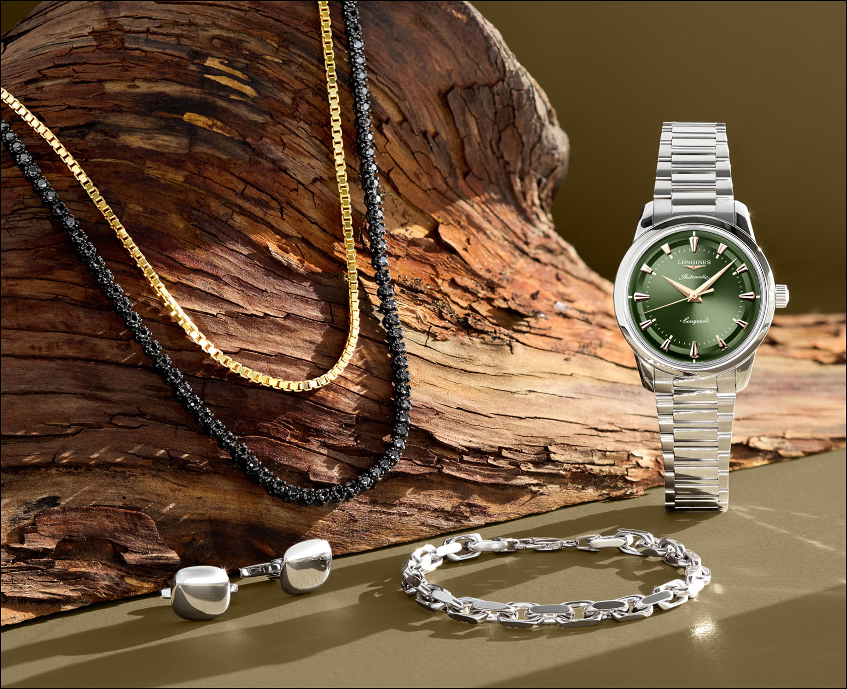 A yellow gold and black diamond necklace are draped over a driftwood log while a stainless steel sports watch with a green dial stands upright next to a silver bracelet.