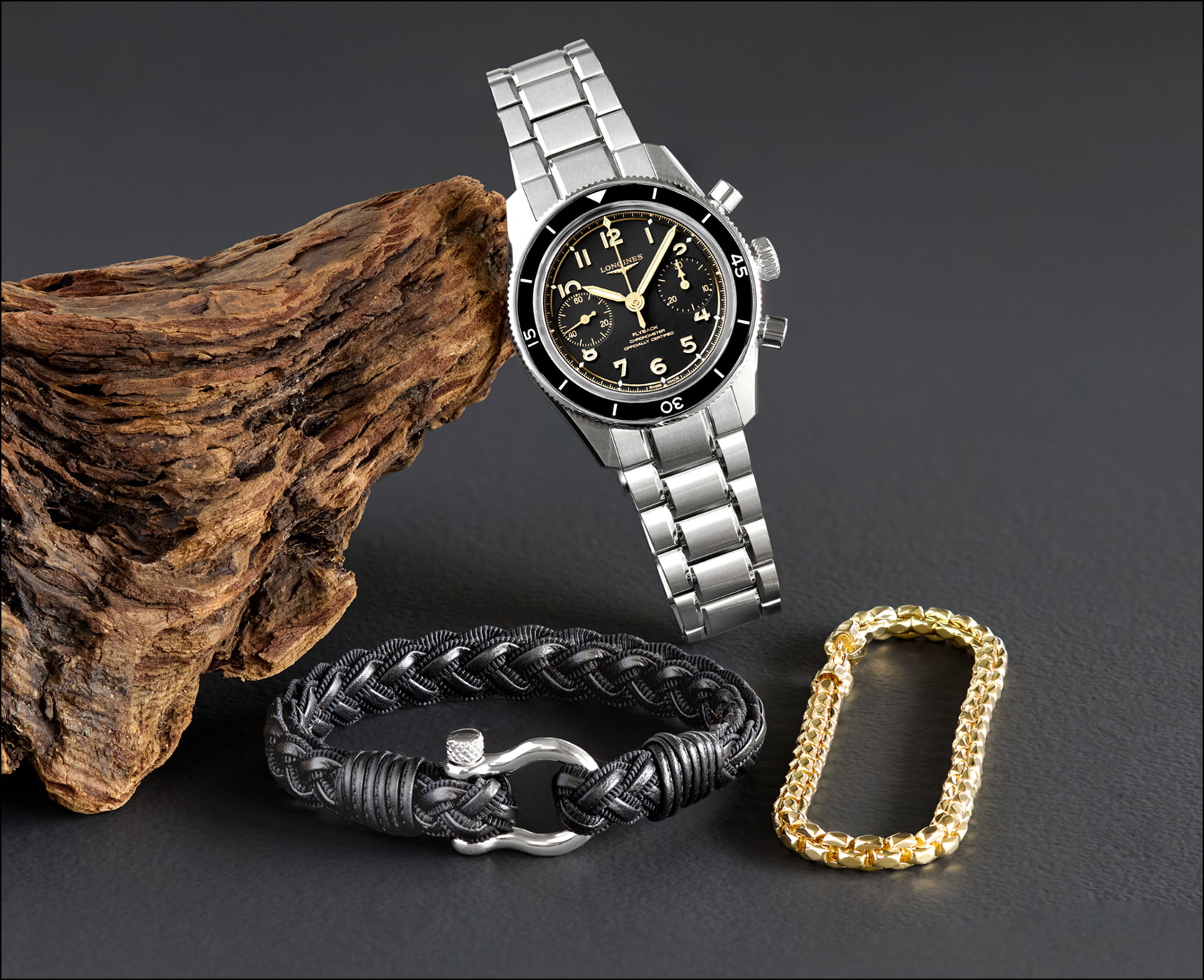 A Longines watch leans against a piece of driftwood next to a black leather bracelet and yellow gold bracelet.